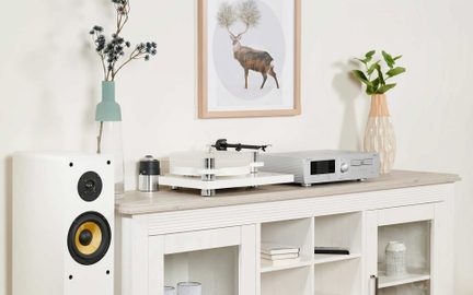 Pohľad spredu na domácu audio súpravu pozostávajúcu z bieleho akrylového gramofónu, strieborného zosilňovača s rebrovaným čelom a bieleho stĺpového reproduktora so žltým kužeľom, umiestnených na svetlom drevenom príborníku s niekoľkými vázami a obrazom na stene v pozadí