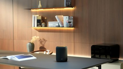 Zwarte draadloze Bluetooth-luidspreker op een designbureau, met houten wandplanken en LED-verlichting op de achtergrond