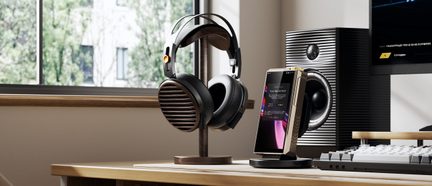 Auriculares hi-fi circumaurales negros sobre soporte de madera, junto a un reproductor de audio digital dorado sobre un escritorio moderno