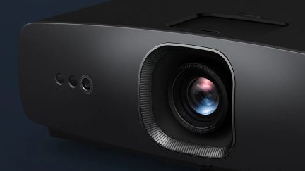 Proyector negro con objetivo central circular, borde texturizado, 3 aberturas laterales redondas, superficie lisa, vista en ángulo