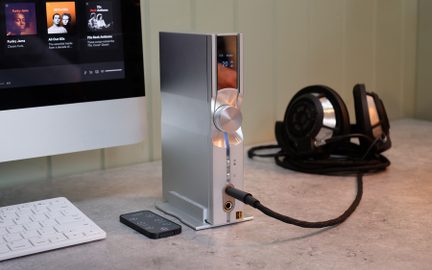 Stříbrný vertikální audio zesilovač s kruhovým tlačítkem, digitálním displejem, připojeným kabelem, černými sluchátky, dálkovým ovladačem a bílou klávesnicí