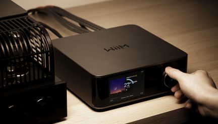 Amplificador de áudio WiiM preto com ecrã a cores na frente e botão rotativo, vista em três quartos sobre móvel de madeira