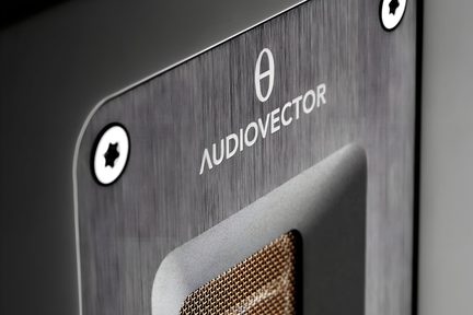 Skråt nærbillede af frontpladen i grå børstet metal på en lydenhed, prydet med « Audiovector »-logoet, to stjerneskruer og en rektangulær åbning med gyldent gitter