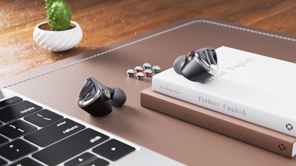 To trådløse in-ear-høretelefoner i metallisk antracitgrå med sølvfarvede kanter, trekantet gitter på fronten og sorte silikonepropper, set oppefra på et skrivebord