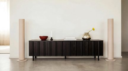 Zwart laag dressoir met geribbelde deuren, minimalistische decoratie met een rookglazen vaas, een kaars en twee beige zuilen aan elke kant