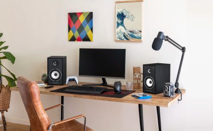 Wooden desk with two black speakers, a computer monitor, a keyboard, a mouse, and two colorful paintings on the wall