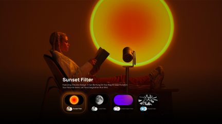 Sidevisning af en cylindrisk projektor i grå metal på stativ, der projicerer en bred, cirkulær, orange-gul solnedgangslignende lysring på en mørk baggrund, med en kvinde siddende i profil i forgrunden, læsende en bog.