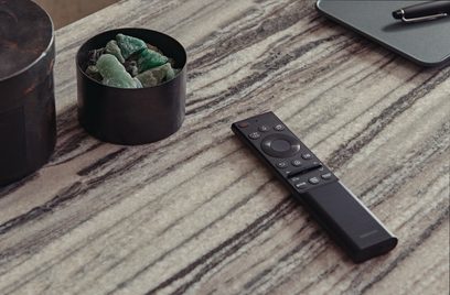 Černá dálkový ovladač Samsung, hnědý keramický hrnek s tmavě zelenými kameny na světlé dřevěné desce s žilkováním