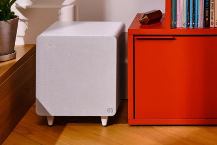 Bílý textilní subwoofer v krychlovém provedení na nožkách, v obývacím pokoji vedle červeného nábytku