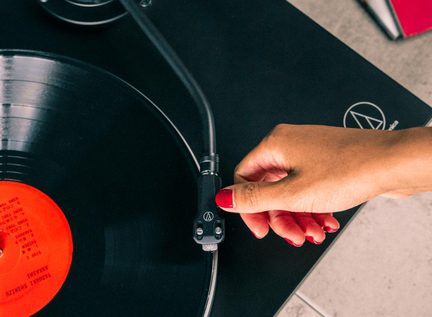 Tæt nærbillede fra oven af en sort pladespiller, tonearm holdt af en hånd, sort vinylplade med rød etiket