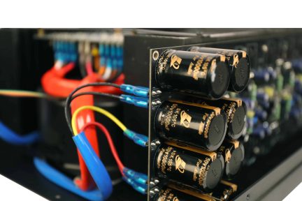 Lähikuva elektronisen laitteen sisäosista: rivissä mustia, kultamerkittyjä sylinterimäisiä kondensaattoreita kiinnitettynä metalliselle piirilevylle, ympärillä monivärisiä, suojattuja johtonippuja