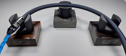Dwa ciemnobrązowe drewniane podstawki na kable audio z prostokątną podstawą i czarną metalową łukowatą konstrukcją, z podłączonym niebieskim kablem