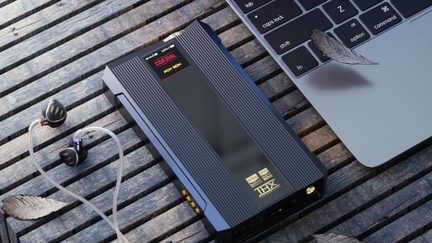 Ampli-DAC portátil negro metálico con acabado acanalado, banda central brillante y pequeña pantalla que indica «COAXIAL», conector de auriculares dorado visible en el frontal, vista en ligera perspectiva tres cuartos sobre listones de madera, con auriculares intrauditivos de cable translúcido y esquina de teclado de ordenador portátil.
