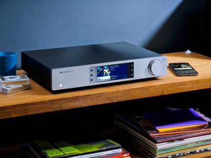 Amplificatore audio Cambridge, vista frontale, posizionato su uno scaffale di legno, con telecomando, smartphone e libri nelle vicinanze