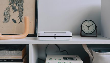 bílá police s černým rámem, béžová svíčka, bílé elektronické zařízení, kulaté černé hodiny, složené knihy, košík z látky