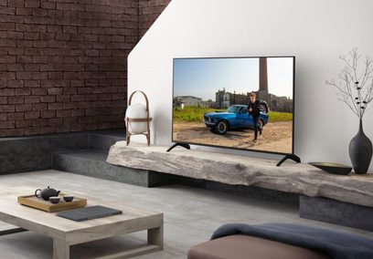 Televizor Panasonic TX-49HX940E zobrazující modré auto zaparkované před cihlovou budovou, umístěný na kamenném nábytku v moderním interiéru
