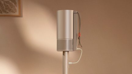 Metal silindirik bir cihazın yan görünümü, delikli ızgara, tekstil kordon, turuncu düğme, beyaz kablo, bej arka plan