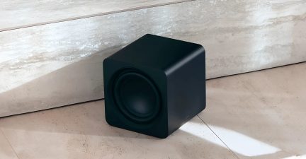 Černý subwoofer se zaobleným krychlovým designem, pohled z tříčtvrtečního úhlu na světlé podlaze u kamenné zdi