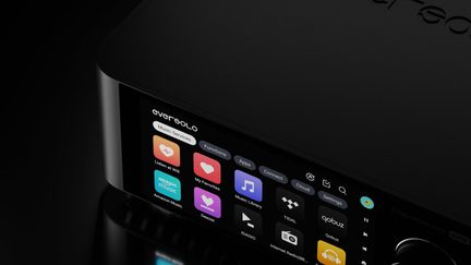 Detailní záběr v tříčtvrtečním pohledu na černou hliníkovou obdélníkovou audio jednotku, jejíž přední panel dominuje barevný dotykový displej zobrazující ikony hudebních aplikací, doplněný vpravo kovovým otočným knoflíkem.