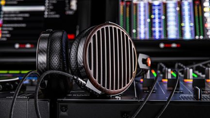 Over-ear hovedtelefoner i sort og brun med åbne træskaller og store lodrette metalgitter, perforerede læderørepuder og sort fletet kabel, set i tæt sidebillede placeret på et mixerbord