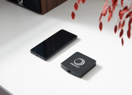 Černý obdélníkový smartphone s vypnutým displejem a černé čtvercové zařízení s logem Elipson na bílé ploše