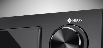Sort drejeknap til lydstyrke med hvidt "HEOS"-logo på mørkegrå frontpanel, tæt forfra-udsigt