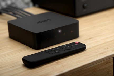 Matně černý plastový multimediální box s přední LED a ikonami, černý dálkový ovladač vpředu, pohled z 3/4 na dřevěném stole