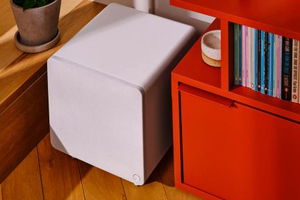 Bílý subwoofer s minimalistickým designem, umístěný na podlaze vedle červené knihovny