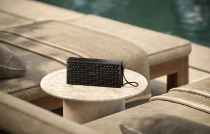 Altoparlante portatile Loewe nero con griglia traforata e cinturino da polso, su un tavolo rotondo beige a bordo piscina