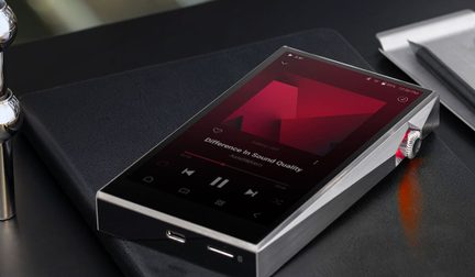 Lettore audio digitale in metallo spazzolato con schermo tattile, pulsante rotativo, visualizzazione della musica, poggiato su un taccuino nero