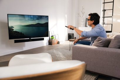 Mand sidder på en grå sofa i en stue og peger med en fjernbetjening mod et stort vægmonteret fladskærms-tv, der viser et maritimt landskab