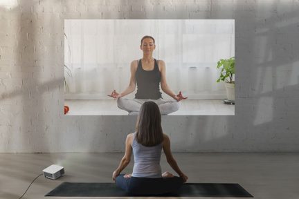 Kvinde set bagfra på sort yogamåtte, vendt mod en projicering af en mediterende person, hvid murstensvæg, hvid projektor på gulvet
