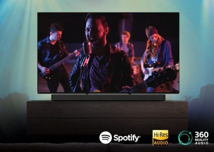 Plokščiaekranis televizorius ant medinio baldo, muzikantų grupė fone, apačioje Spotify, Hi-Res Audio, 360 Reality Audio logotipai.