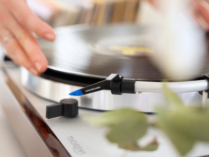 Levá ruka nastavující vinylovou desku na gramofon s kazetou Concorde Music a bílým ramenem pro čtení
