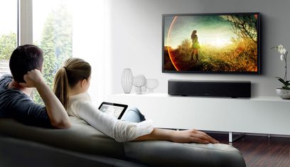 Yamaha YSP-5600 soundbar under et fjernsyn, der viser en kvinde på en mark, et par sidder på en sofa og ser på hende