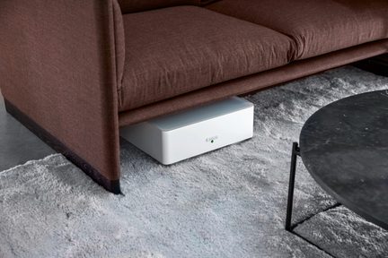 Płaskie, dyskretne białe urządzenie elektroniczne pod brązową kanapą na szarym dywanie, minimalistyczny design do ukrytego przechowywania