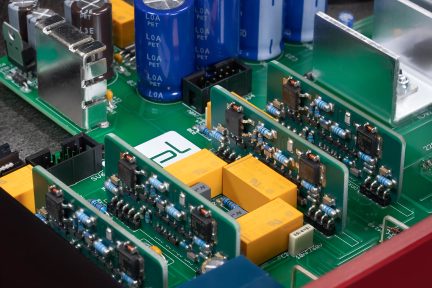 Primo piano di una scheda elettronica verde con condensatori blu, relè gialli e moduli di circuiti integrati verticali