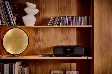 Sort Roberts-radio med centralt display og to højttalere på en hylde nær en rund lampe og cd'er