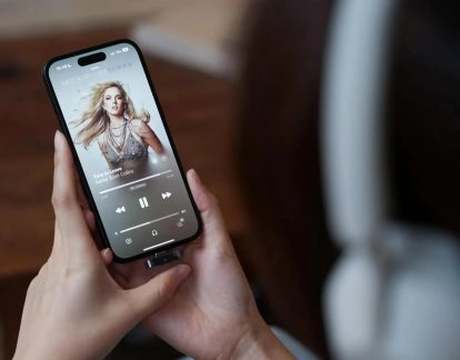 iPhone negru ținut cu ambele mâini, cu player muzical pe ecran și un mic adaptor conectat la portul inferior